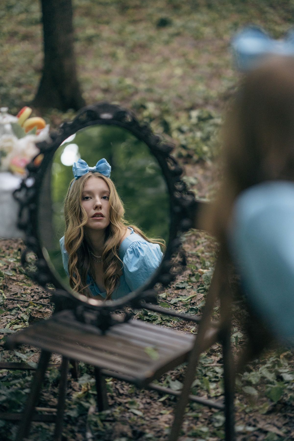 Young woman dressed as Alice in Wonderland reflected in ornate mirror outdoors in forest setting.