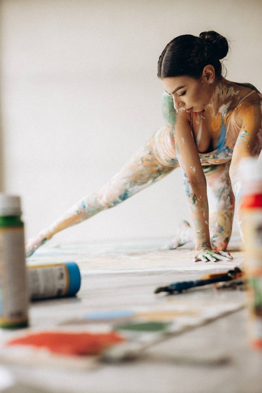 A paint-covered female artist kneels on the floor, working on a large canvas surrounded by colorful brushes and paint tubes.
