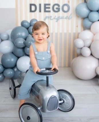 Toddler boy named Diego Miguel sits on a vintage toy car surrounded by blue and white balloon garlands.