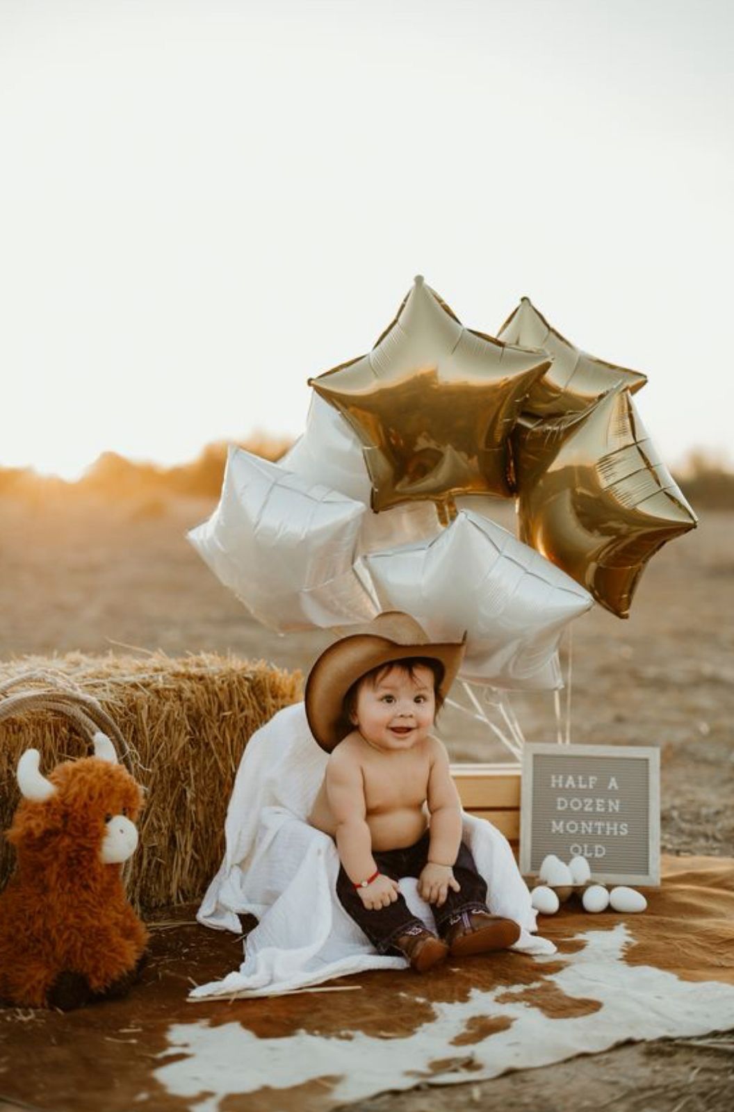 Baby in cowboy hat sits by hay bale with gold star balloons and a half dozen months old sign.