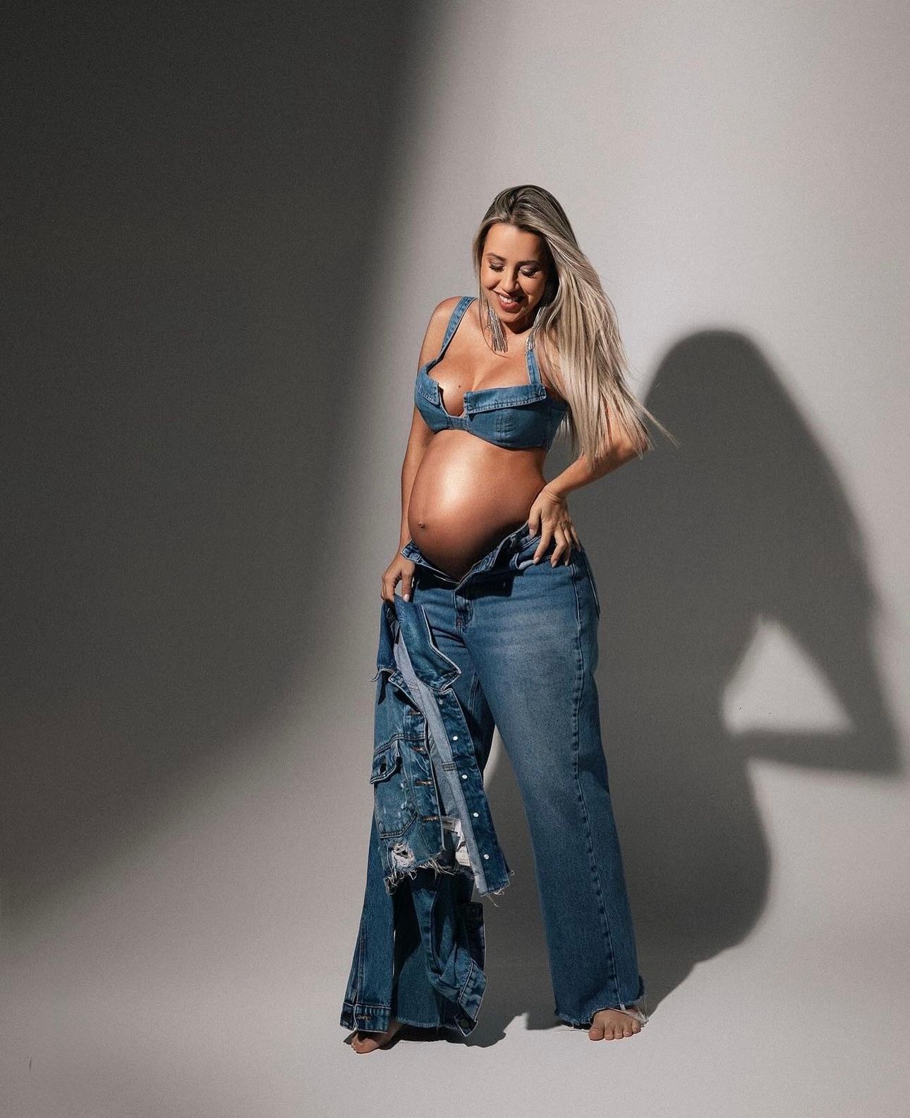Pregnant woman in denim bikini top and wide-leg jeans showing baby bump during maternity photoshoot.