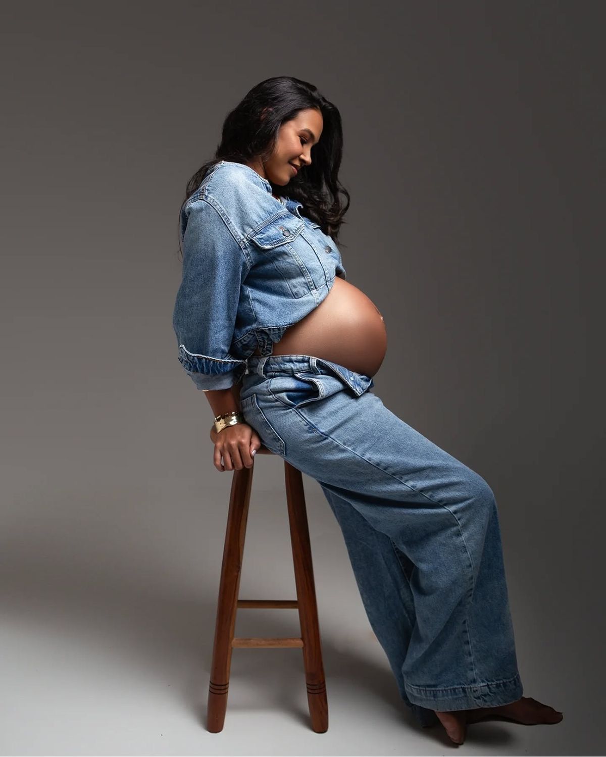 Pregnant woman in denim jacket and wide-leg jeans sitting on a wooden stool, cradling her baby bump in a studio.