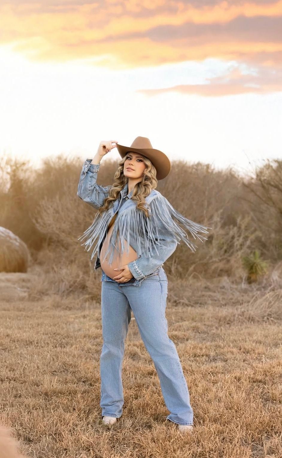 Woman in fringed denim jacket, cowboy hat, and jeans poses in a field at golden hour sunset.