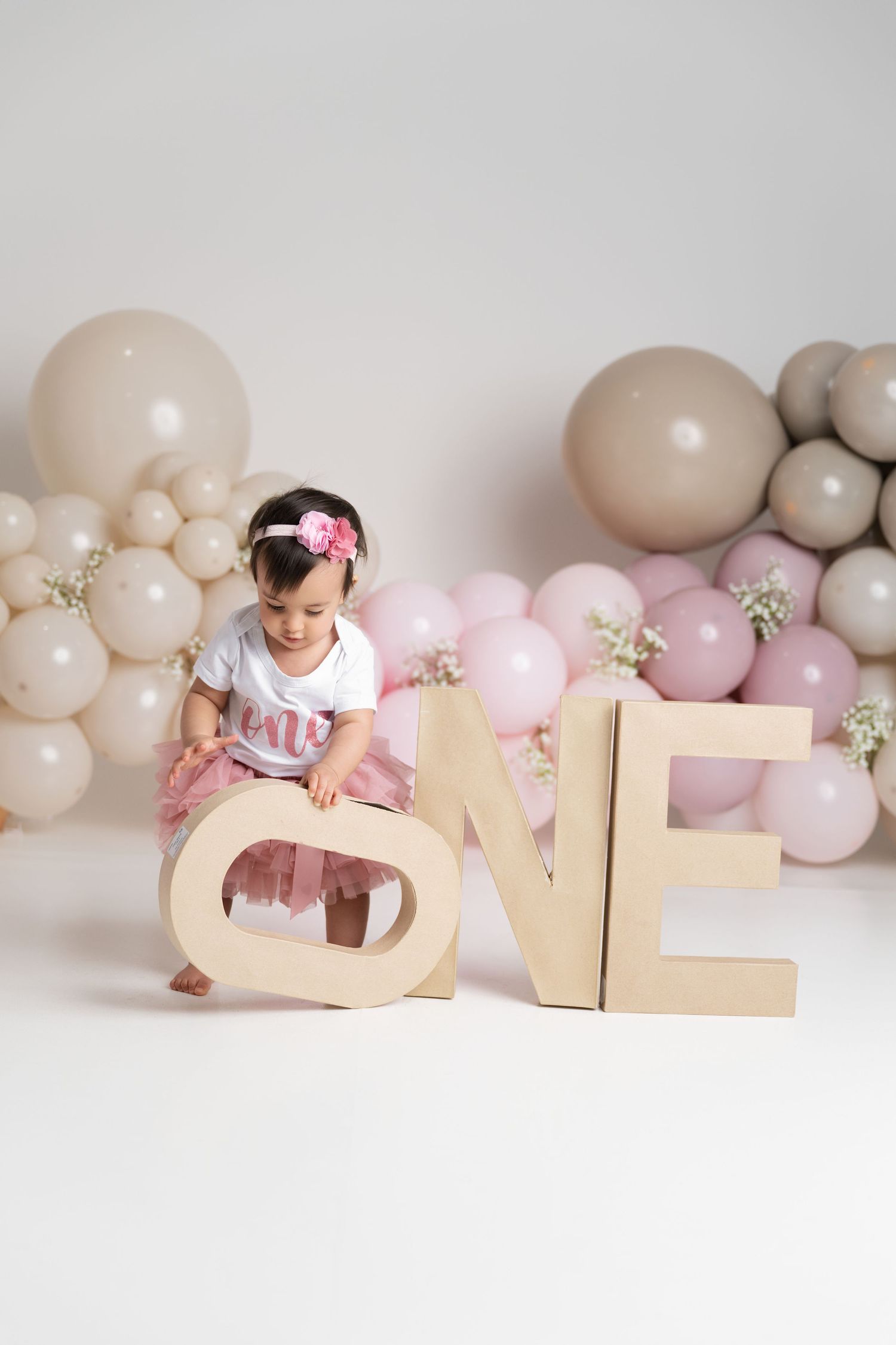 A baby girl in a pink tutu sits between large wooden ONE letters surrounded by beige and pink balloons for her first birthday.