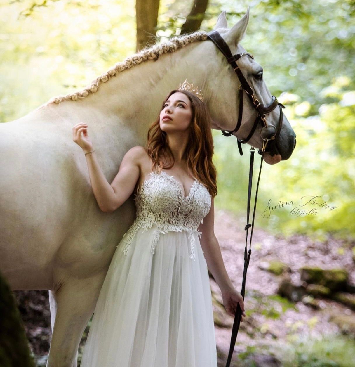 A bride in a white lace gown stands beside a white horse in an enchanted forest setting.