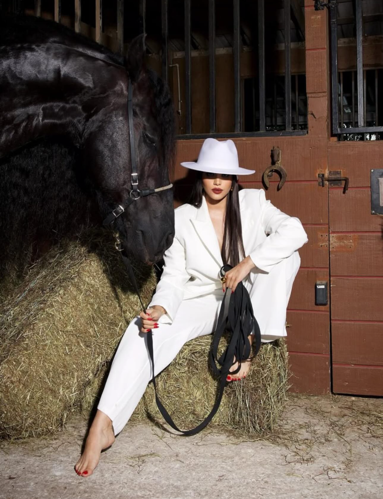 Woman in white suit and hat poses barefoot with black horse on hay in stable setting.