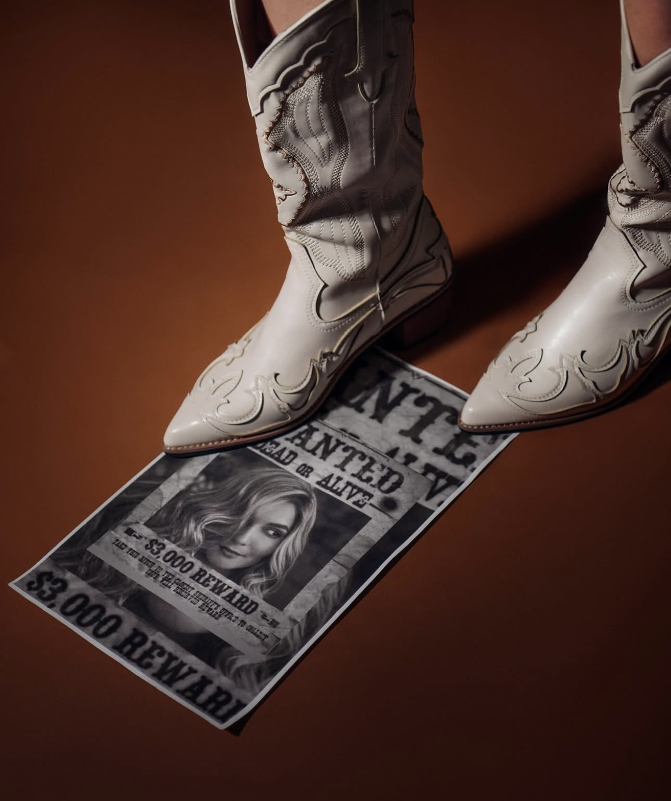 White cowboy boots standing on a Western wanted poster with $5,000 reward on brown background.