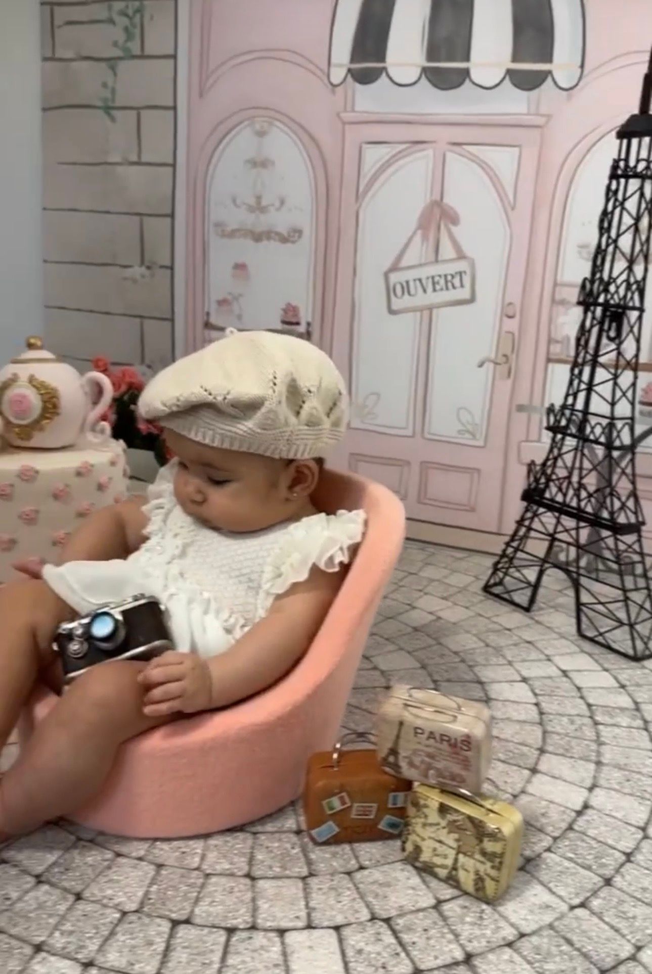 Baby in beret hat sits in pink chair with mini Eiffel Tower, Paris suitcases, and camera in French-themed photo shoot.