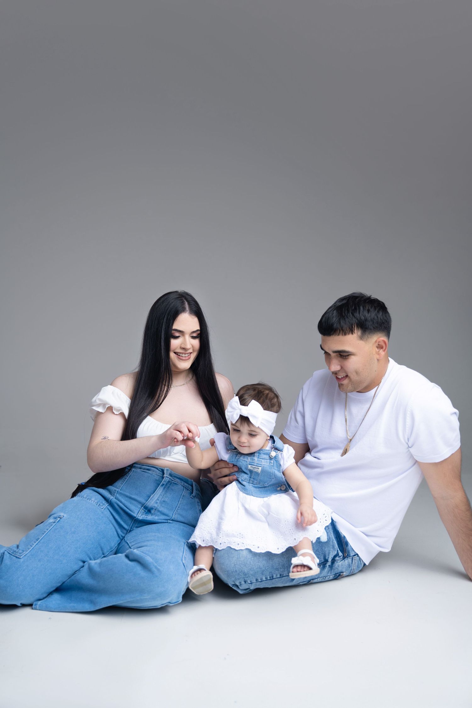 Family in matching casual denim and white outfits sitting together on gray backdrop for studio portrait.