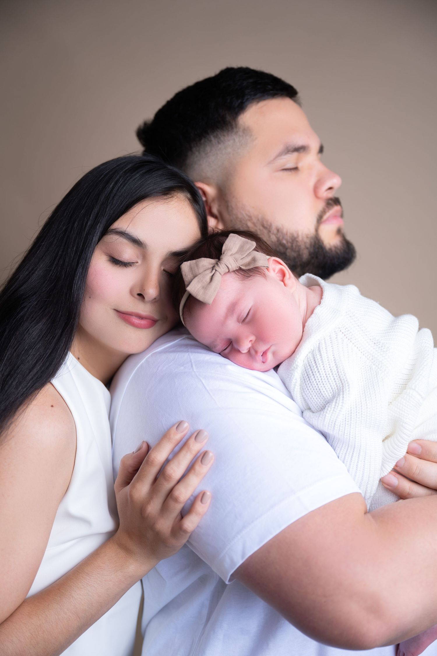 A sleeping baby wearing a pink bow rests peacefully on someone's shoulder in a tender family portrait.