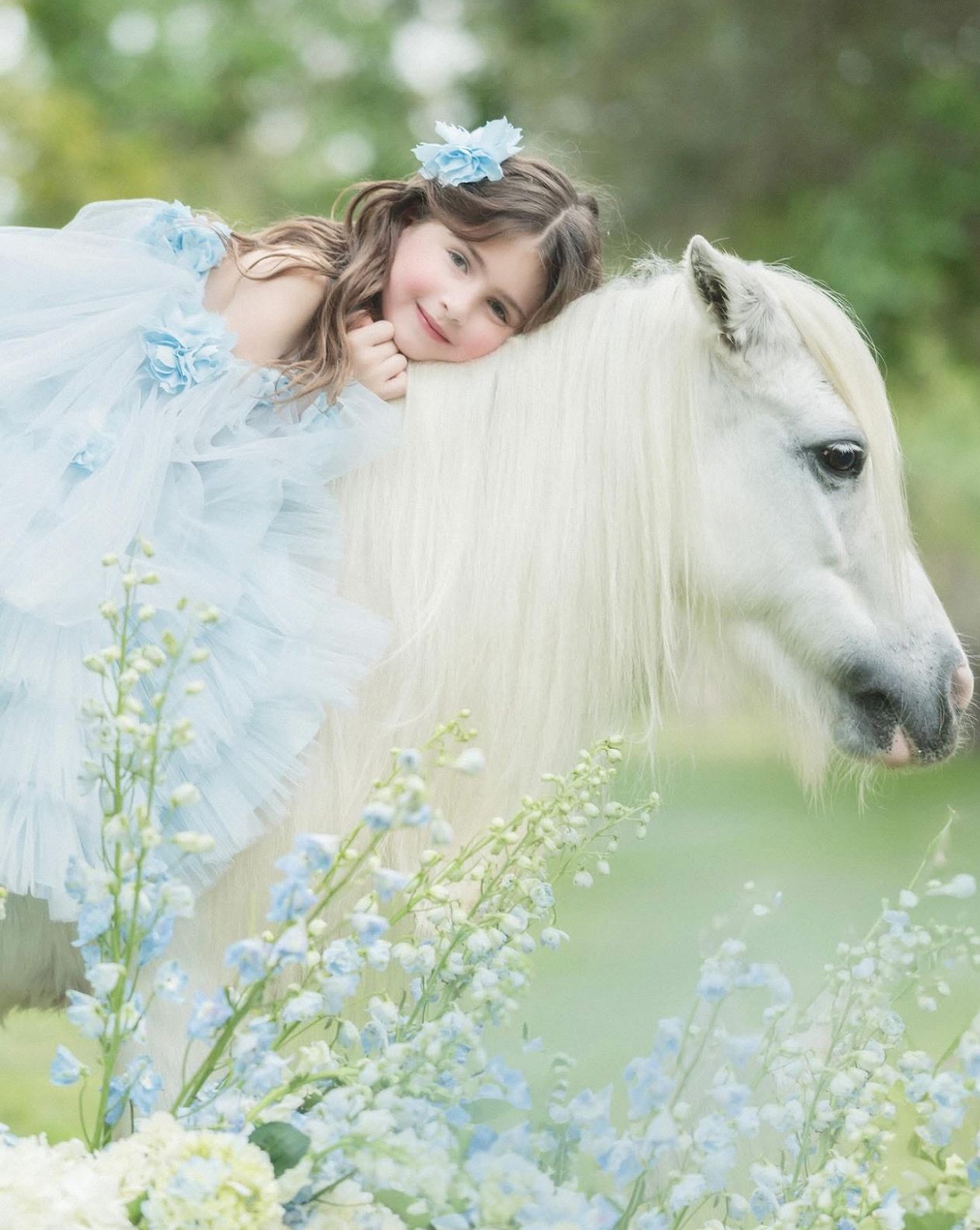 A young girl in a blue tulle dress leans on a white pony surrounded by delicate blue flowers in a dreamy garden.