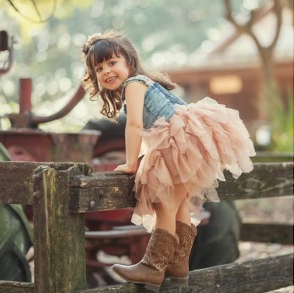 Young girl in denim top, pink tutu, and cowboy boots sitting on a rustic wooden fence at a farm.