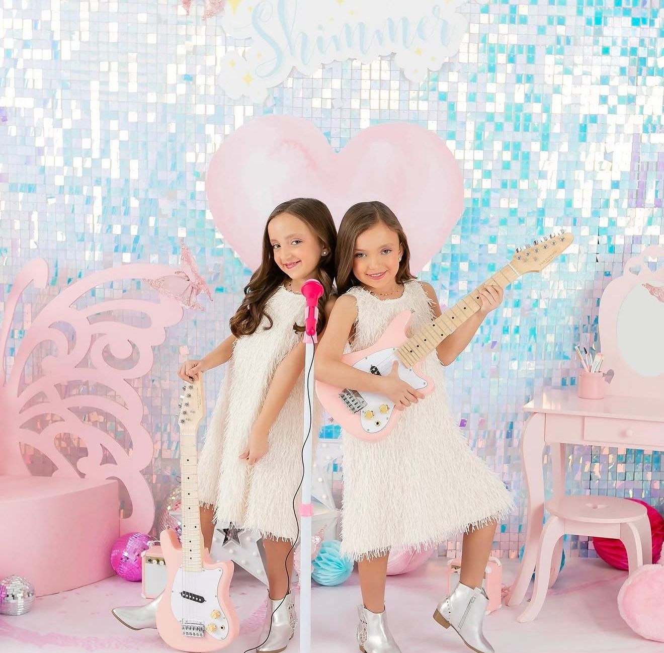 Two young girls in white dresses pose with a microphone and guitar against a sparkly iridescent backdrop.