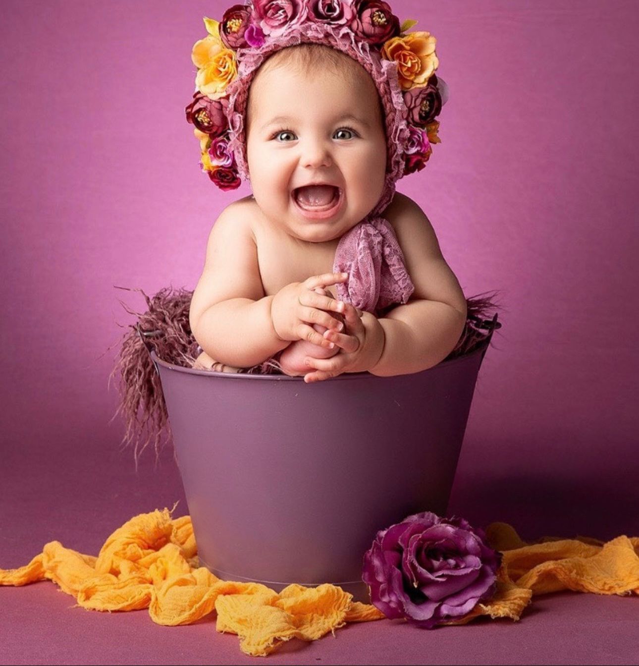 Joyful baby wearing floral crown sits in purple pot with yellow and purple roses against pink background.