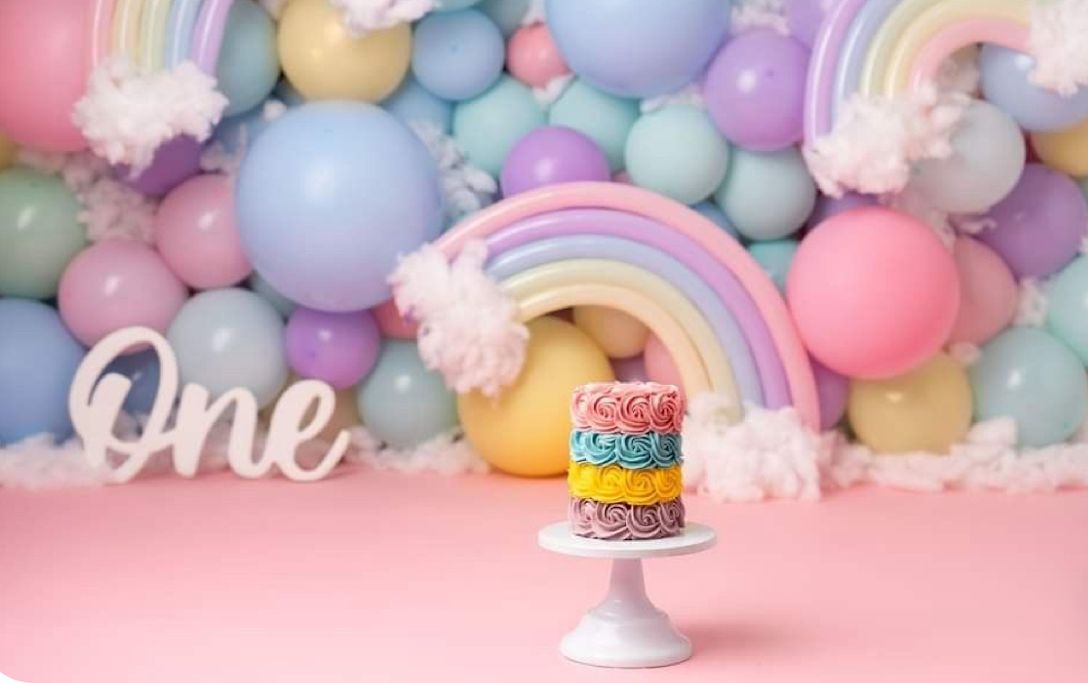 Colorful rainbow birthday cake on white pedestal with pastel balloon arch and 'One' script text decoration.