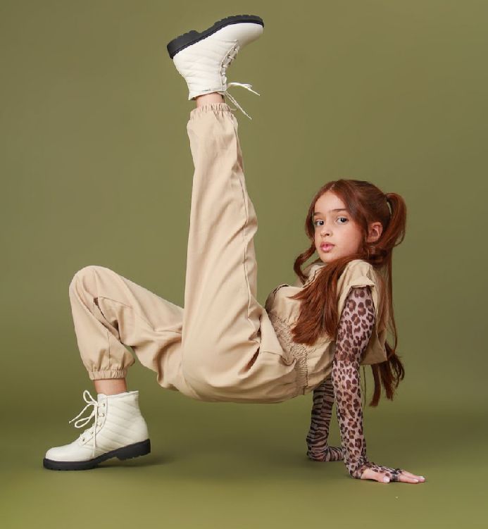 Fashion model in beige cargo pants and white boots strikes dynamic pose against olive green backdrop.