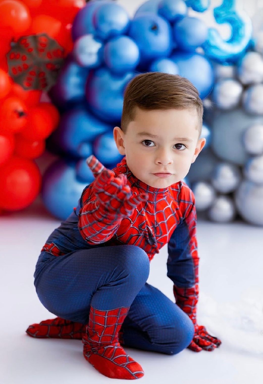 A child in a Spider-Man themed outfit crouches against a backdrop of red, white and blue balloon decorations.