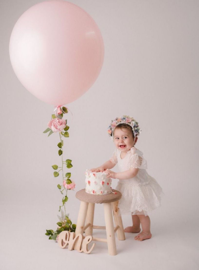 First birthday celebration setup with pink balloon, floral decorations, white dress, and small cake on wooden stool.