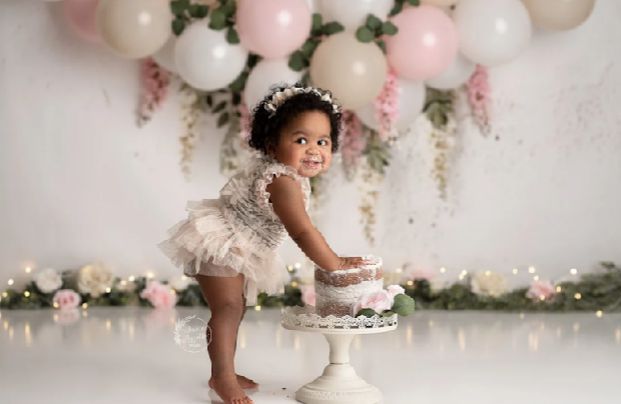 A baby in a frilly white dress sits on a vintage cake stand surrounded by pink and white balloons and floral decor.