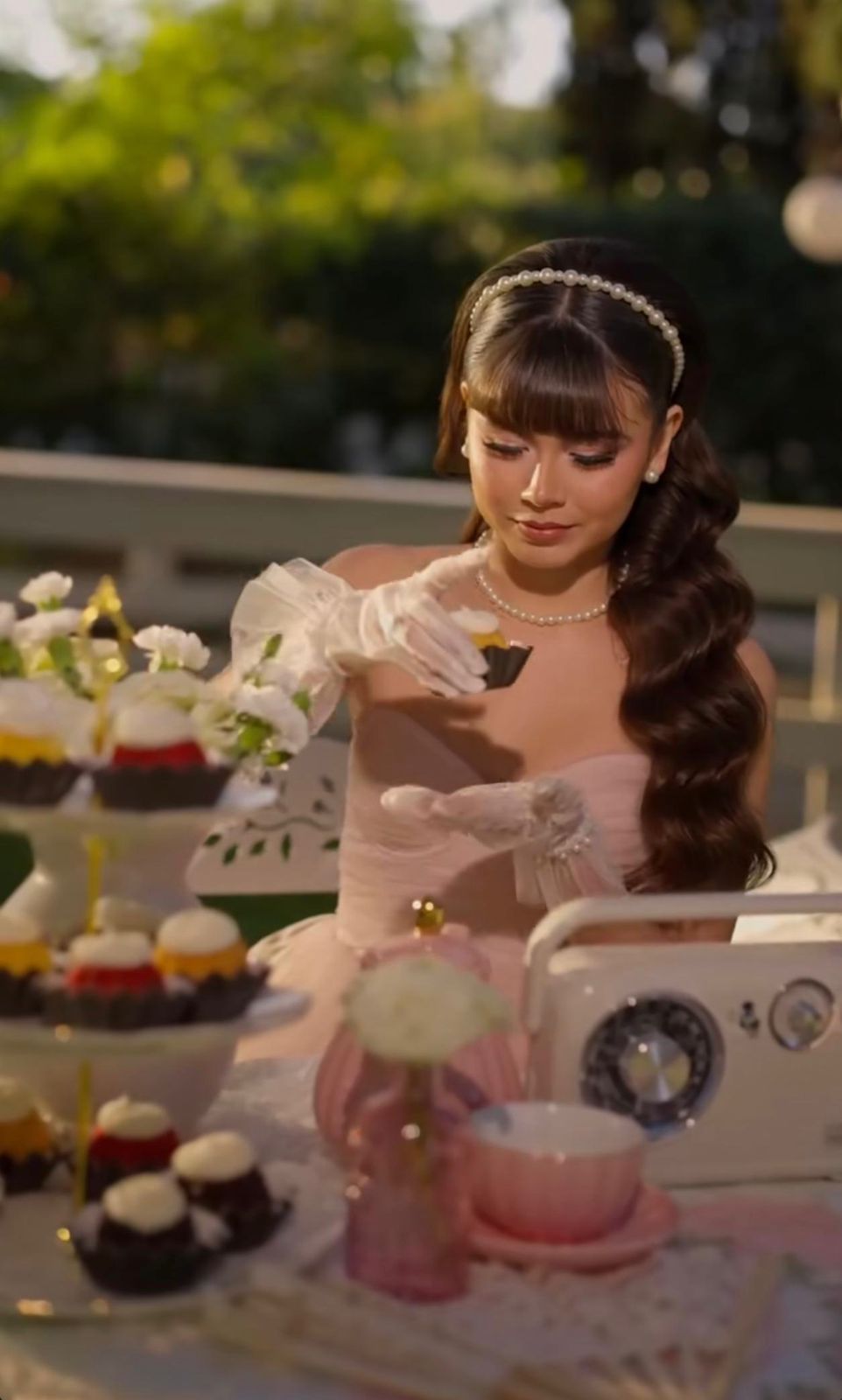 Elegant woman in pink dress with pearl accessories arranging cupcakes at an outdoor dessert table.