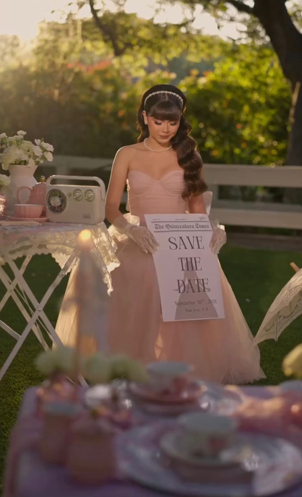 Woman in pink gown holds Save the Date sign at elegant outdoor garden party with vintage decor.