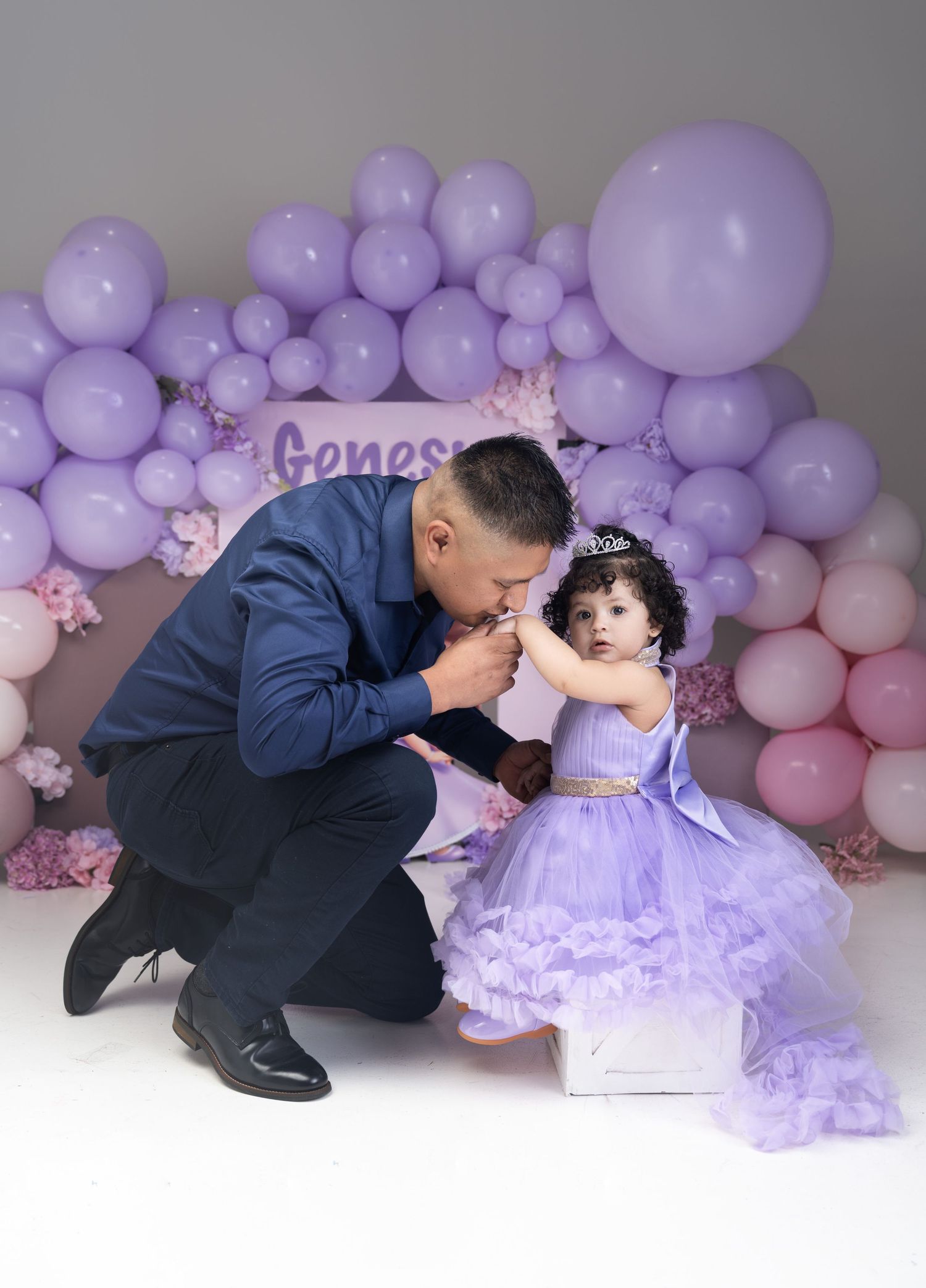 A father kisses his baby girl's hand at her first birthday party with purple balloons and floral backdrop.