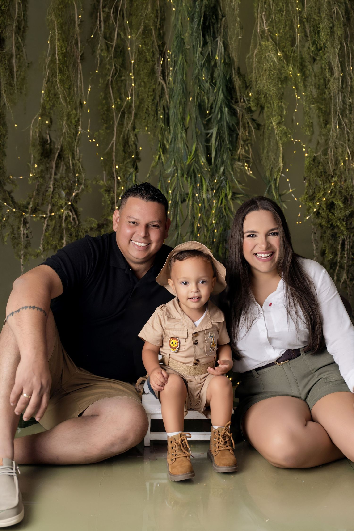 Family portrait with parents and baby boy in safari outfits, sitting before hanging greenery with fairy lights.