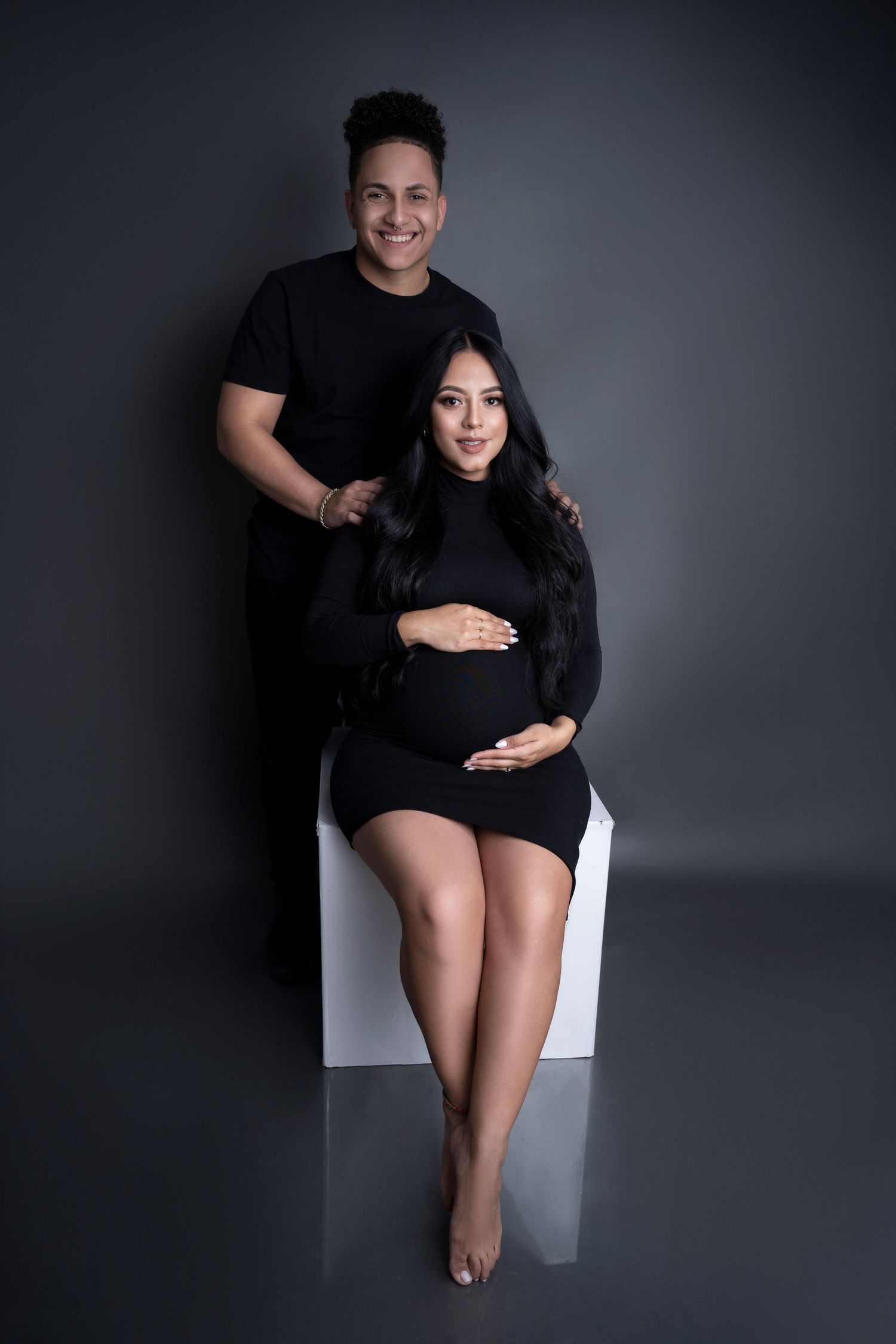 A pregnant woman in a black dress sits while her partner stands behind her, both smiling in a studio maternity photo.
