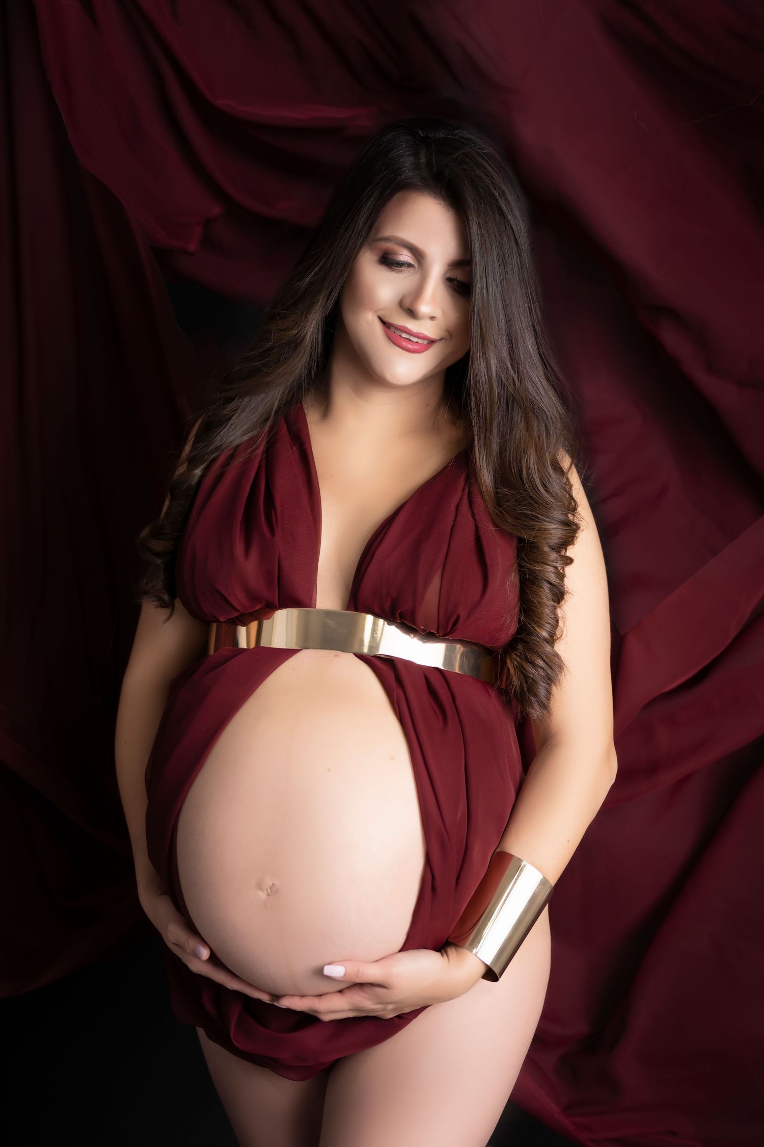 Smiling pregnant woman in a burgundy halter dress with gold belt and cuff, cradling her bare baby bump against draped fabric.
