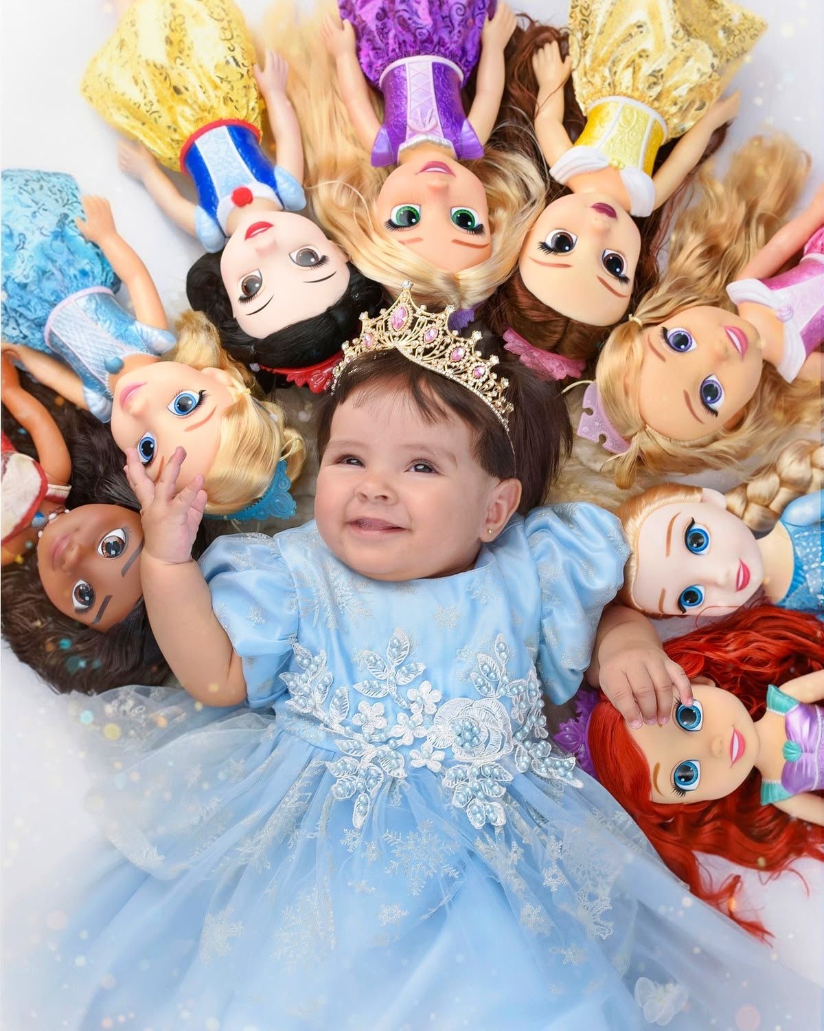 Baby girl in blue Cinderella dress wearing tiara, surrounded by Disney princess dolls, lying on white background.