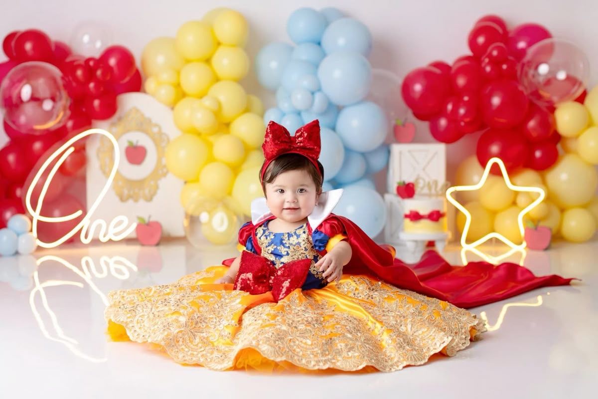 Baby girl in Snow White dress sits before colorful balloon backdrop at first birthday cake smash photo shoot.