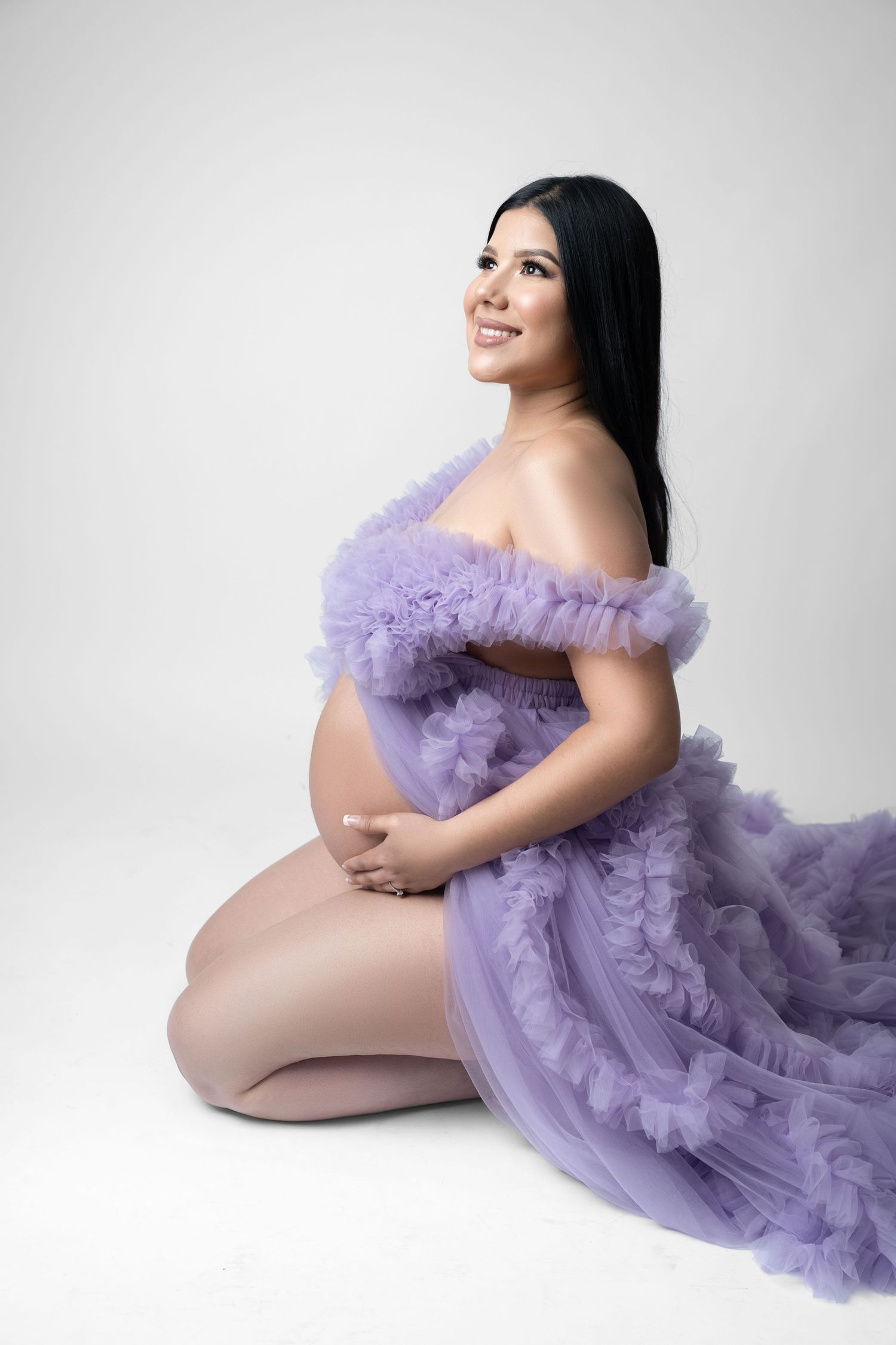 Smiling pregnant woman kneeling in elegant purple ruffle tulle maternity gown against white background.