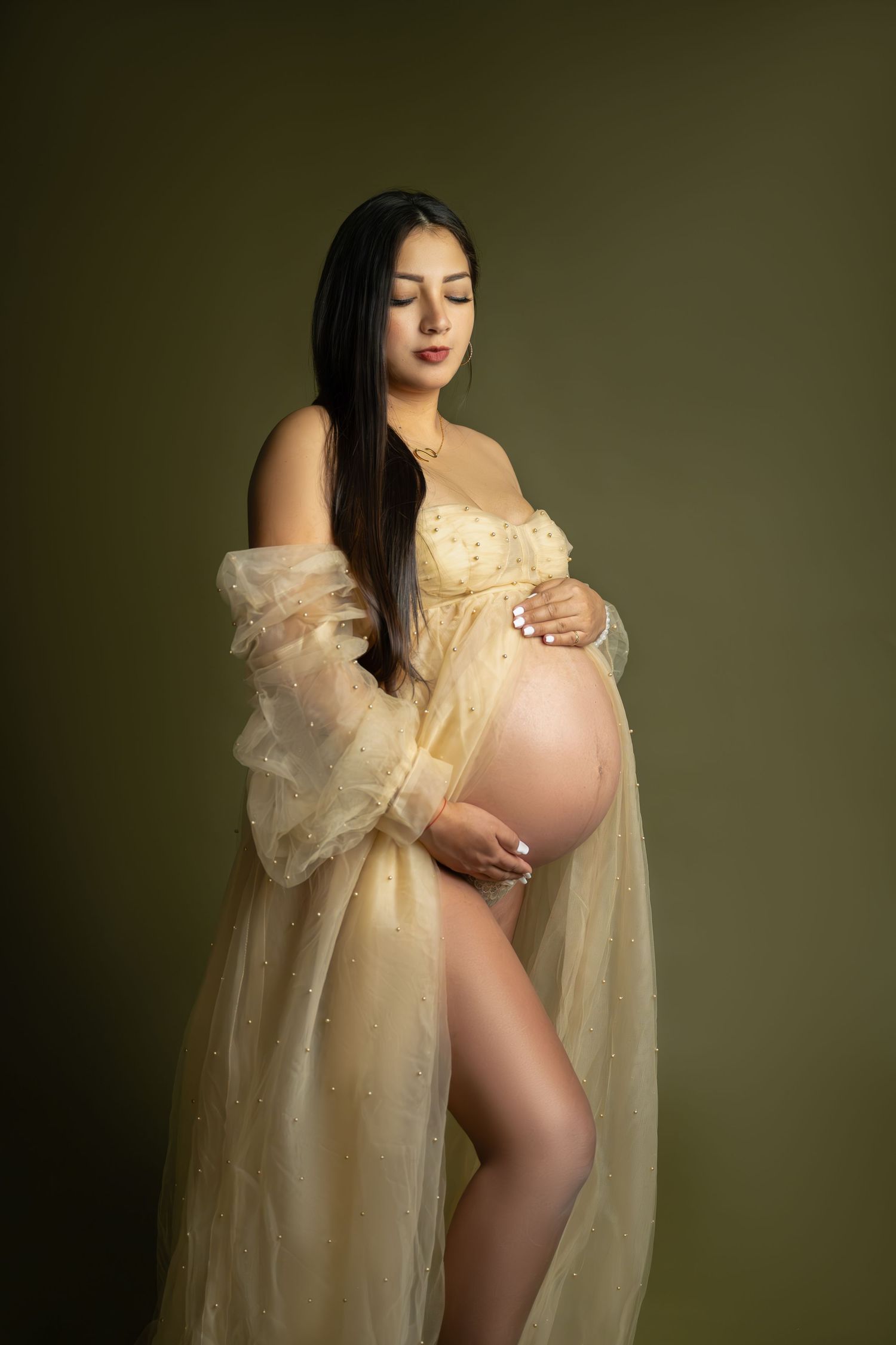Pregnant woman in cream lingerie and sheer robe cradling her baby bump against an olive green background.