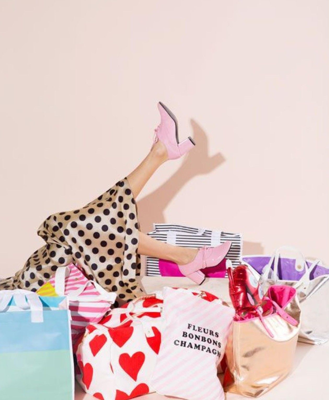 Person buried in colorful shopping bags wearing pink heels and polka dot dress, with gift bags and heart print wrapping paper.