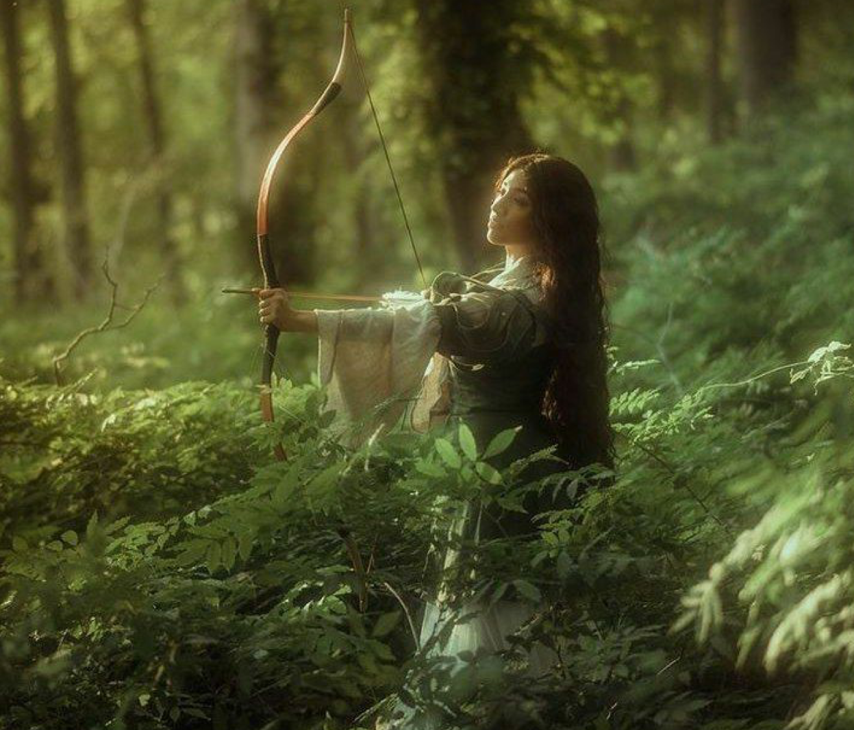 A woman archer in a forest draws a bow among lush green ferns, dressed in medieval fantasy attire.