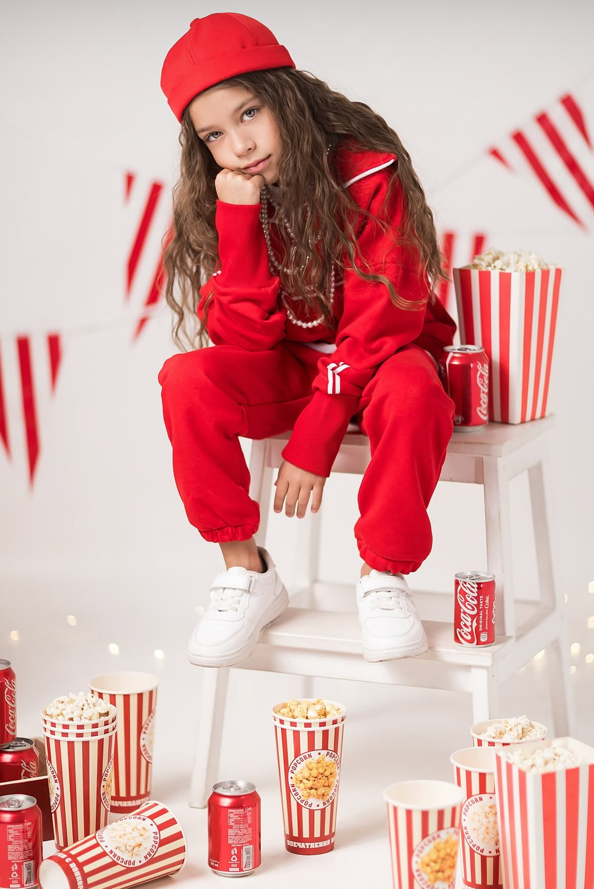 A young girl in a red tracksuit and cap sits surrounded by Coca-Cola cans and striped popcorn buckets.