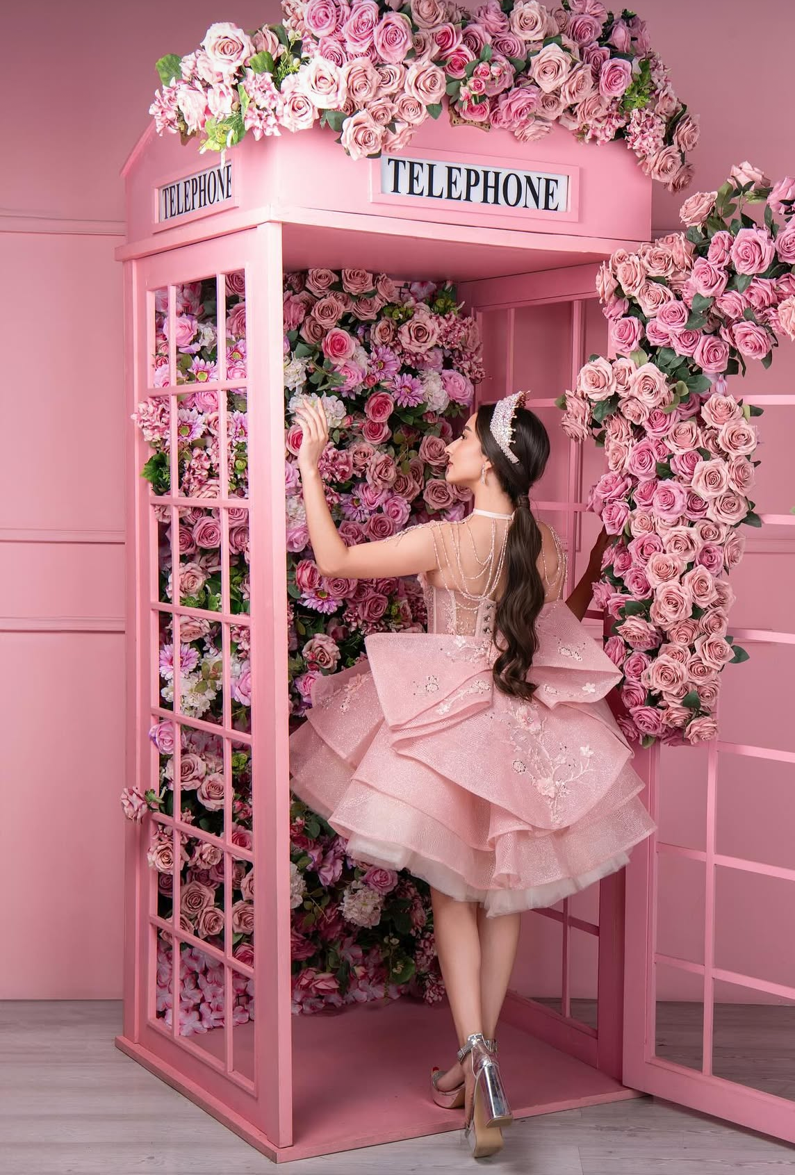 A young woman in a pink ballgown poses inside a pink telephone booth adorned with pink roses.