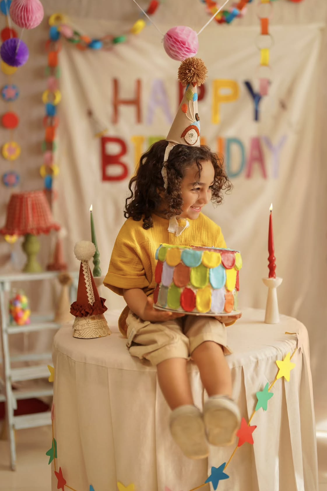 A smiling child in a party hat holds a colorful birthday cake at a festive decorated table with candles and Happy Birthday banner.