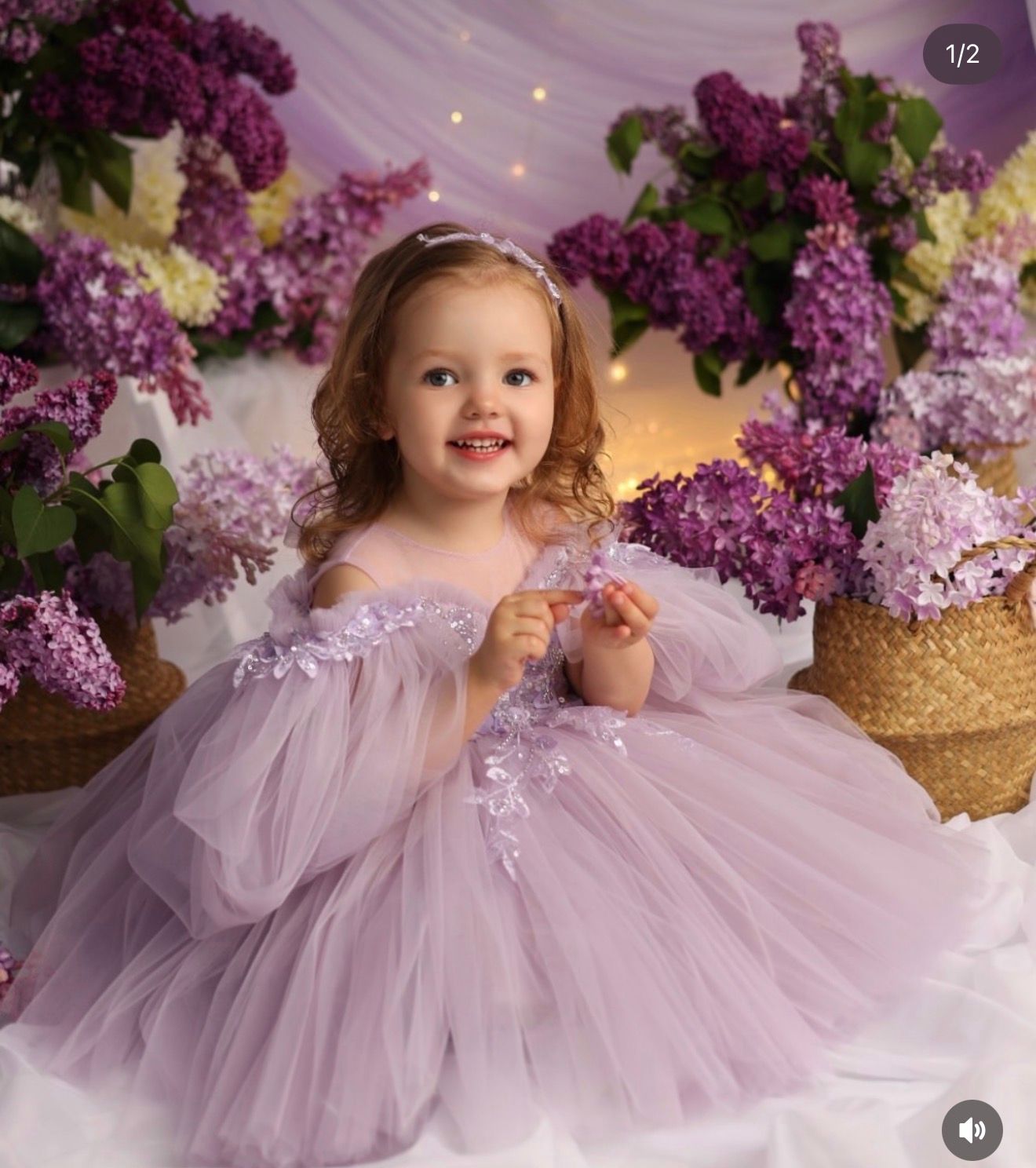 Toddler girl in lavender tulle dress surrounded by purple lilac flowers in a dreamy floral portrait setting.