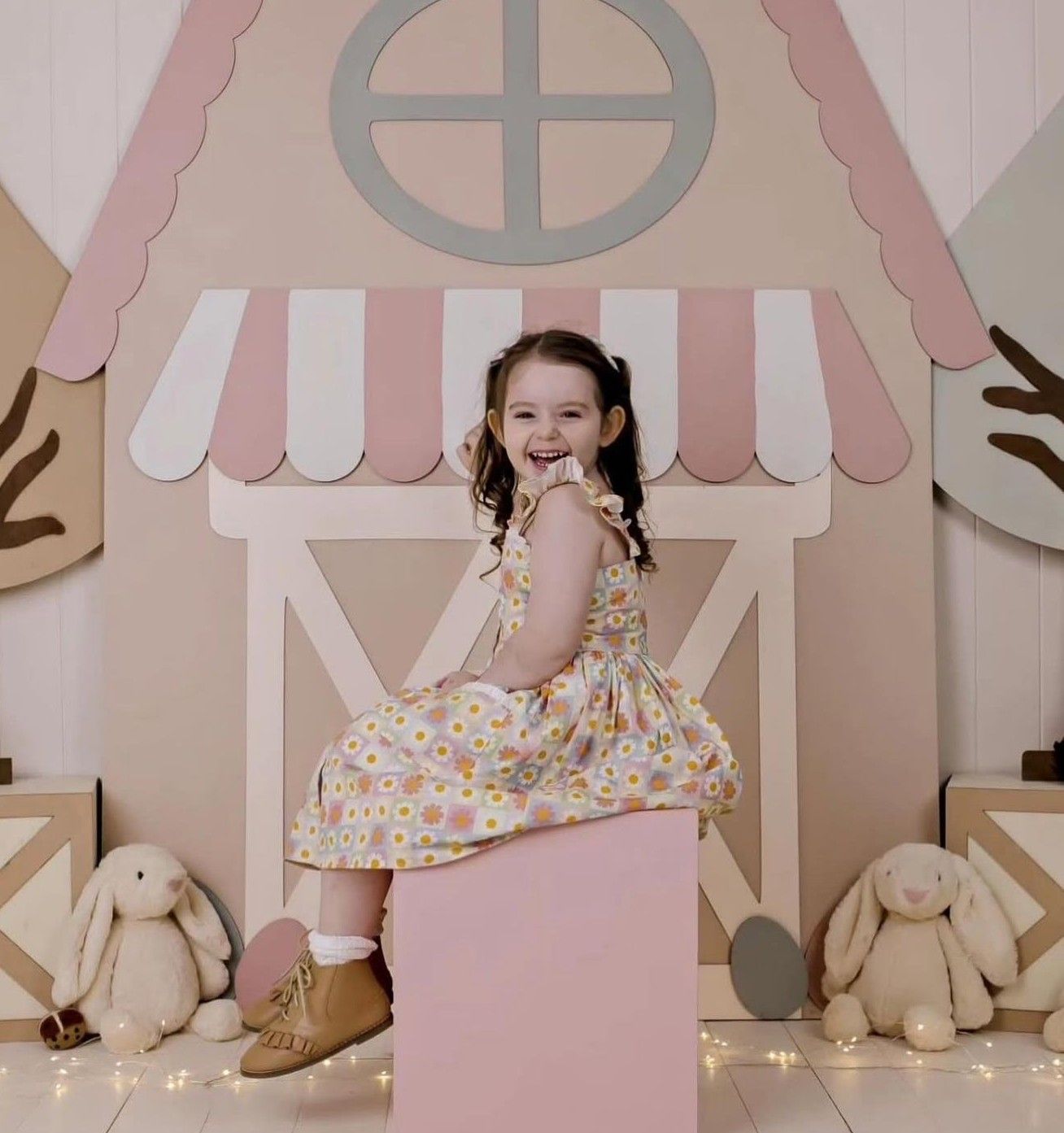 Young girl in floral dress sits smiling on pink block before a pastel toy house backdrop with bunny plush toys.