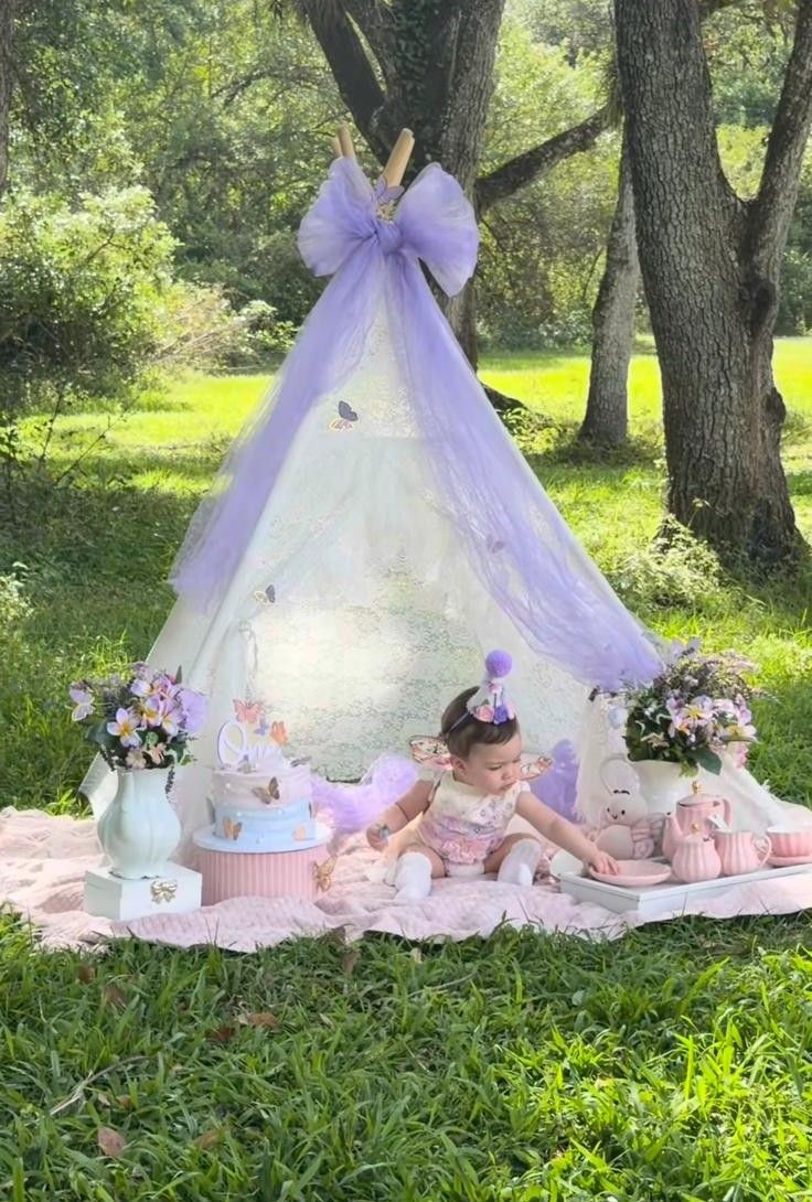 Baby girl in pink dress sits under a lavender tulle teepee with birthday cake and flowers outdoors.