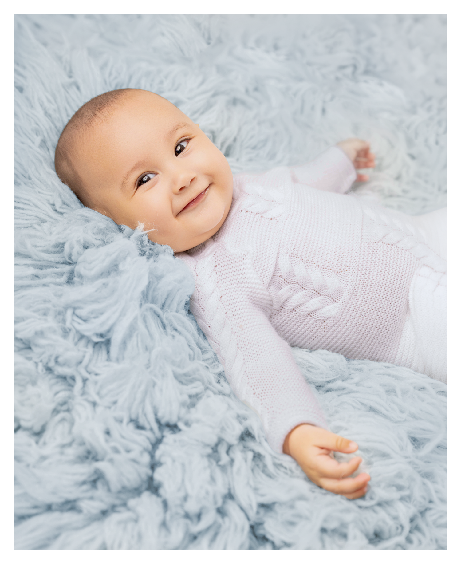 Smiling baby lying on a fluffy light blue blanket wearing a white knit sweater.