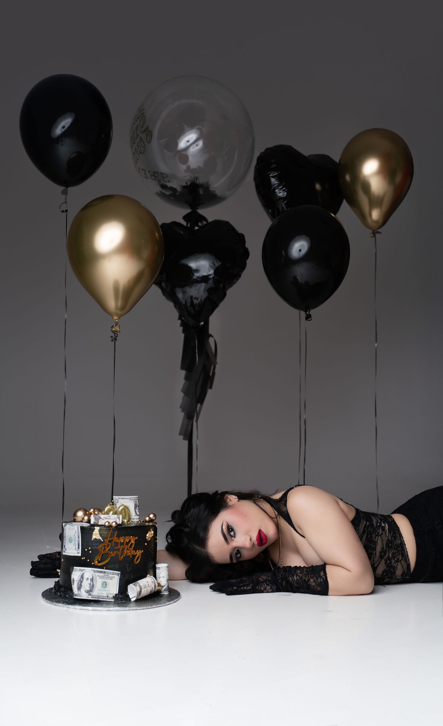 Woman in black lingerie lying beside a birthday cake with gold and black balloons in a moody studio setting.