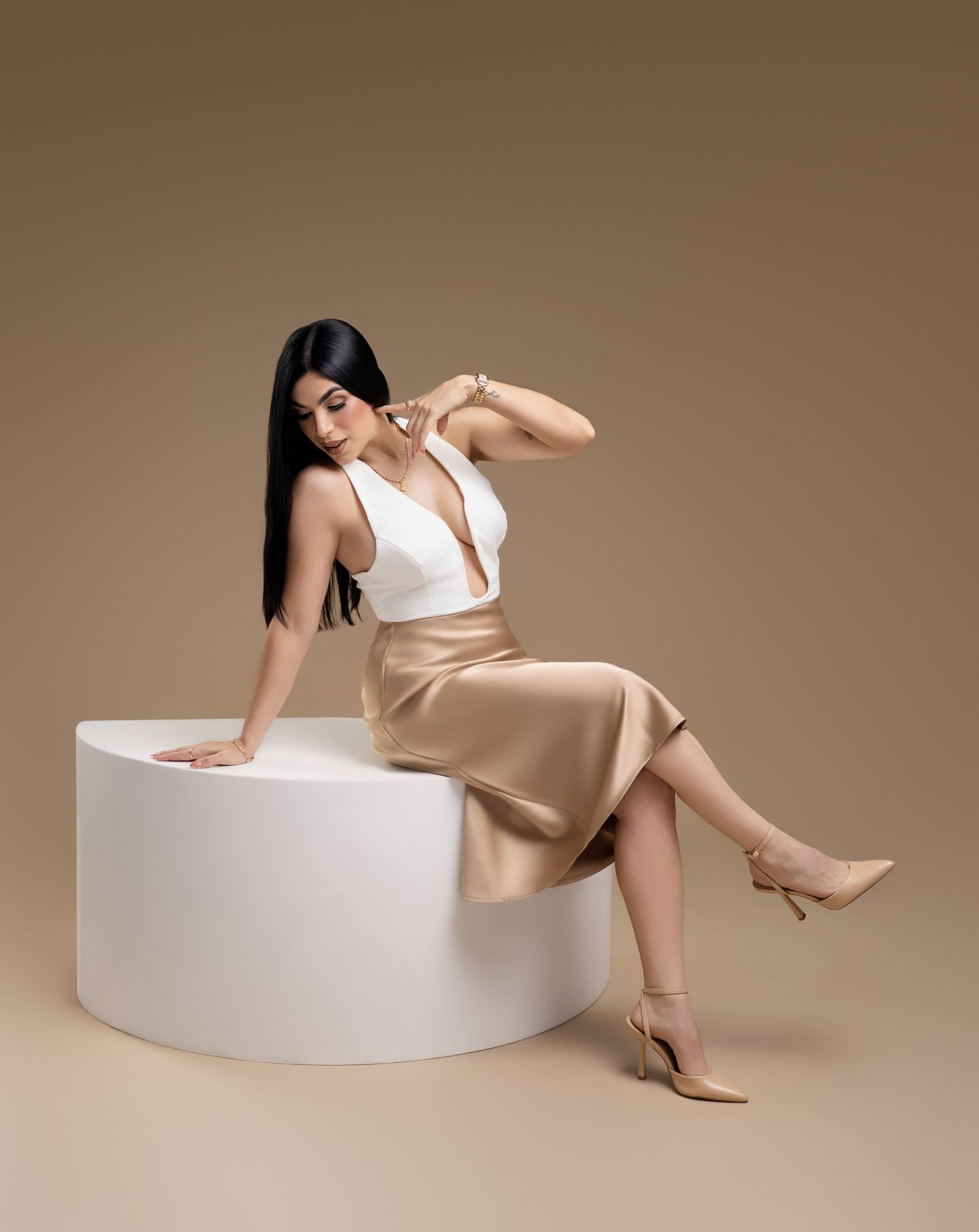 Woman in white top and gold satin midi skirt seated on white pedestal with nude heels against beige background.