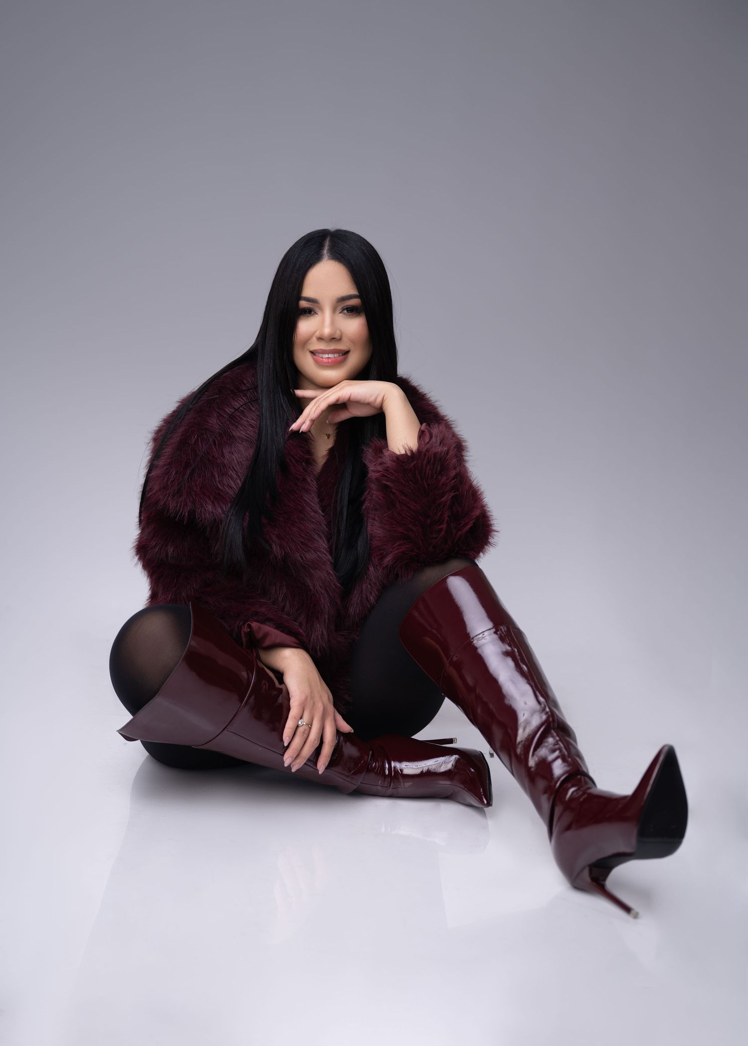 Woman with long black hair smiling on a white studio floor in a burgundy faux fur jacket and glossy knee-high stiletto boots.