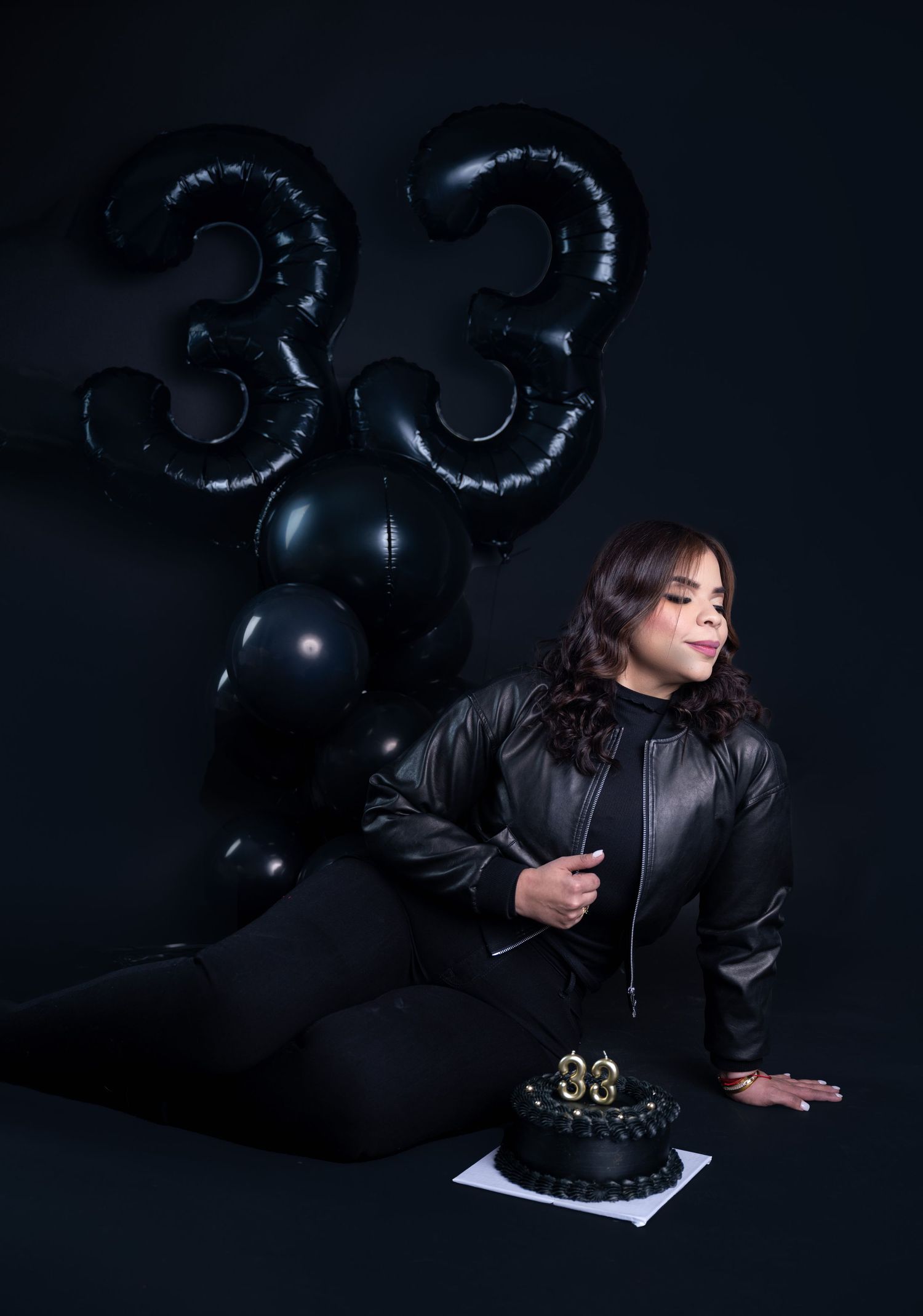 Woman in a black leather jacket sits beside a black 33rd birthday cake with black number 33 balloons in a moody dark photoshoot.
