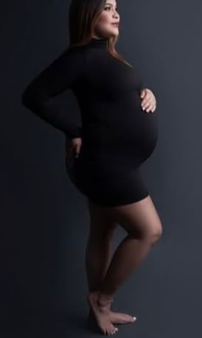 Pregnant woman in a fitted black long-sleeve dress poses barefoot in profile, cradling her baby bump against a dark studio backdrop.