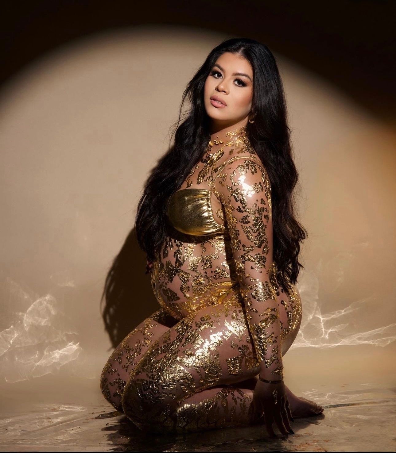 Pregnant woman in gold sequin bodysuit posing gracefully on floor against warm beige backdrop with long black hair.