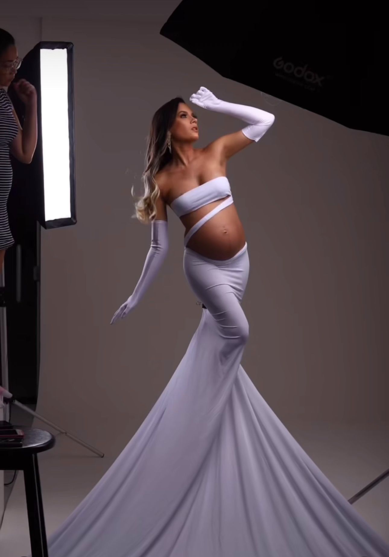 Pregnant woman in white strapless crop top, mermaid skirt, and long gloves poses in a professional photo studio.