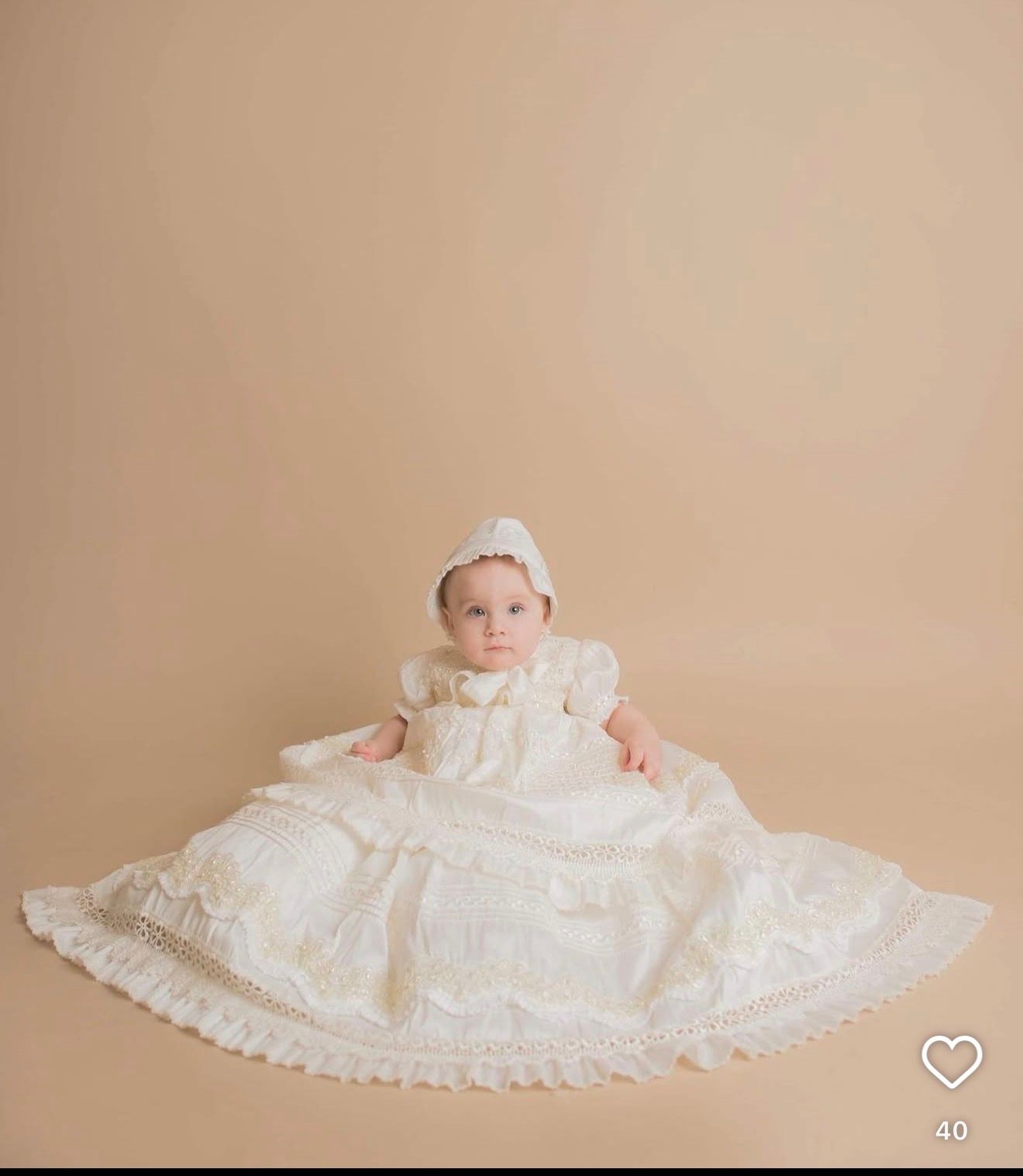 Baby in ivory lace christening gown and bonnet posed on beige backdrop for baptism portrait photo.